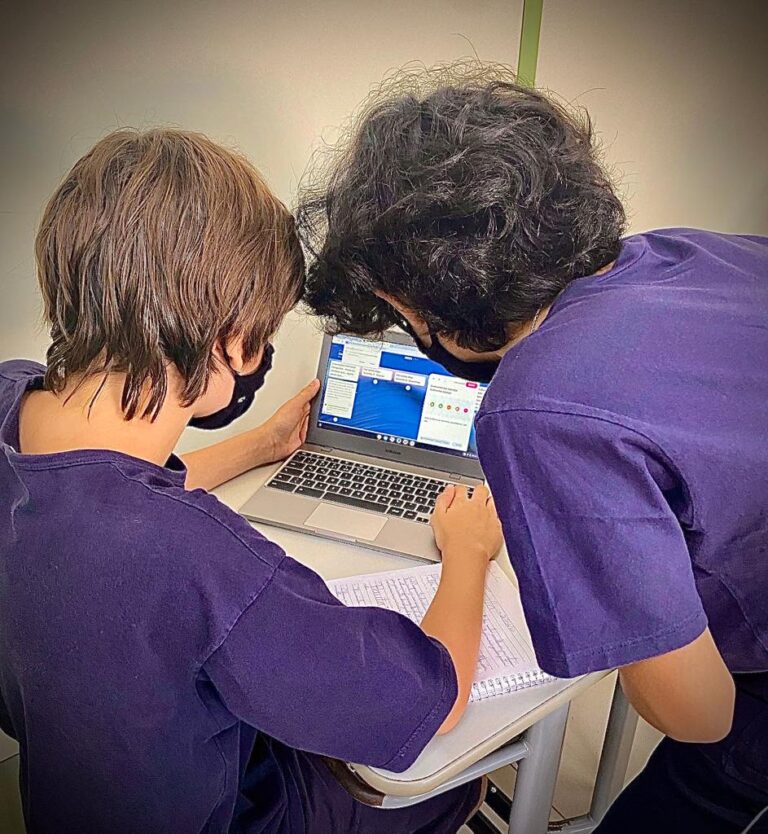 Uma criança está sentada em frente à um laptop, vestindo blusa azul escura e com as mãos nas laterais do equipamento; do seu lado direito está outra criança, vestindo blusa idêntica, encurvada e observando a mesma tela do notebook.